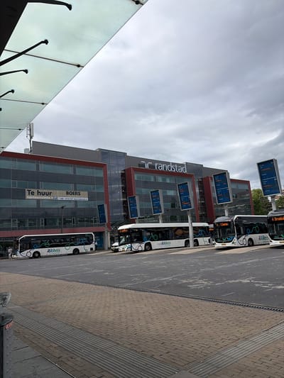 This is a photograph of a bus station in Enschede, Netherlands. The scene is dominated by a modern building with large glass windows, behind which is a dark, cloudy sky. The building has a sign that reads "randstad" in large letters. In front of the building, there are several buses parked. Some of the buses have advertisements for "RRReis" on them. There are also digital display boards showing bus routes and times. The ground is paved with bricks and asphalt. A small number of people are visible, including some standing near the buses and others walking in the distance. The overall impression is that of a functional, urban transportation hub on an overcast day.