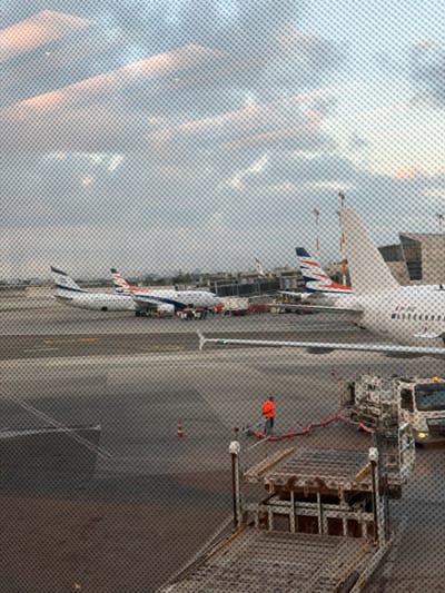 The image depicts an airport tarmac scene, likely in Tel Aviv, Israel, given the presence of El Al airplanes. Two El Al airplanes are visible, one parked at a gate and another further down the tarmac. Several airport vehicles, including baggage carts and service trucks, are present on the ground. A person wearing an orange vest is seen near a baggage loader. The sky is overcast with some clouds. The image appears to be taken through a window with a visible mesh pattern.