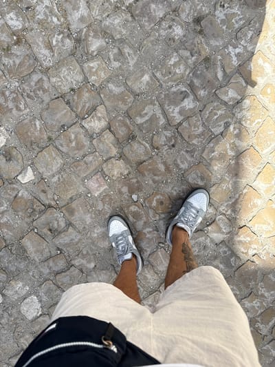 The image shows a person's legs and feet standing on a cobblestone street. The person is wearing white shorts, grey and white sneakers, and a dark fanny pack. The cobblestones are irregular in shape and size, with some cracks and dirt between them. There is a shadow cast on the cobblestones from the right side of the image, suggesting sunlight. The scene is likely a public street in a city. Given the context of Costa da Caparica, Portugal, this could be a street near the coast, or within the town itself.