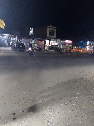 This is a nighttime, outdoor, medium-shot photograph taken in Gampongpaya, Indonesia. The scene depicts a street with several storefronts and food stalls illuminated by artificial lights. In the foreground, the asphalt road is visible, with a sandy, debris-strewn shoulder on the right.
Several people are present. One person is riding a motorcycle in the middle of the road, facing left, with their headlight on. Another person is standing near the motorcycle. Other individuals are visible in front of the shops. A black car is parked on the left side of the road.
Notable details include several signs with text. On the left, "AMRI STYLE" and "AMKI LE" are visible. Further to the right, signs read "KAYA MO", "WM BOUTIQUE", and "WM DIOSC Ngedimsum Premium 2000". A food stall on the right has a sign that says "ES TELER CREAM DURIAN". The setting appears to be a commercial area at night, possibly a market or a street with businesses. The lighting suggests it is dark outside, with the scene illuminated by streetlights and the lights from the businesses. The overall atmosphere is that of a busy, illuminated street at night.