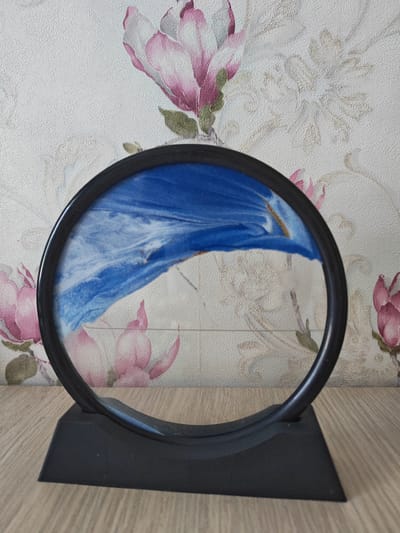 The media file displays a kinetic sand art display in a circular frame with a black base. The sand art itself is abstract, featuring swirls of bright blue and white sand, with hints of gold. The background is wallpaper with a floral pattern, dominated by large pink magnolias and lighter, more abstract floral designs. The texture of the wallpaper is visible, appearing slightly rough.
The kinetic sand art is positioned on a wooden surface, likely a table or shelf, which has a visible wood grain. The lighting appears to be natural, suggesting it is daytime, though no specific time can be determined. The overall impression is one of a decorative item placed indoors, likely in a home or office setting. There are no people visible in the image, nor is there any text. The scene is static, with the sand art in a fixed position. The focus is entirely on the object and its immediate surroundings. The location context of Kogon City, Uzbekistan, is provided as external information and is not visually discernible from the image itself.