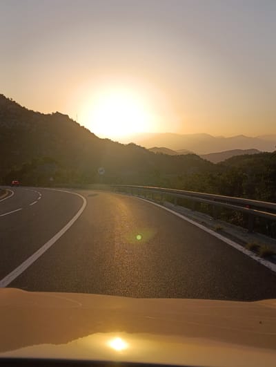 The image captures a winding road at sunset in the vicinity of Gornja Brova, Montenegro. The sun is low on the horizon, casting a warm, golden light across the scene and creating long shadows. The road is paved and marked with white lines, indicating its curvature as it ascends a hillside. A metal guardrail lines the right side of the road. Lush green vegetation covers the slopes of the mountains that frame the road. In the distance, layers of mountains fade into a hazy sky. The foreground shows a portion of a vehicle's hood, reflecting the sunset. A red car is visible on the road in the distance, heading away from the viewer. The overall impression is one of a scenic mountain drive during a beautiful sunset.