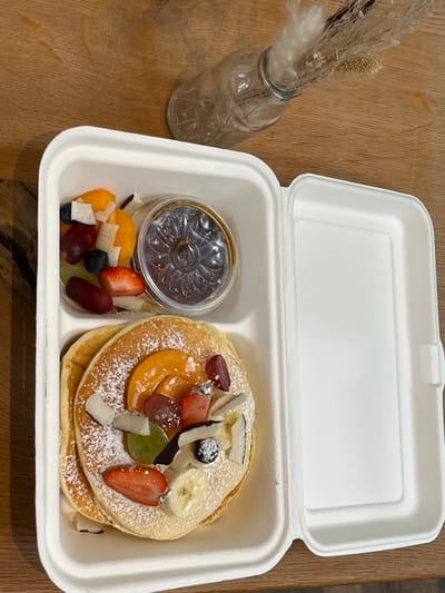 In Esslingen am Neckar, Germany, on a wooden table, a white takeout container holds a stack of three pancakes topped with sliced peaches, strawberries, grapes, bananas, coconut flakes, and powdered sugar. To the left of the pancakes is a small container of syrup. A small bunch of dried flowers in a glass vase is also visible on the table.