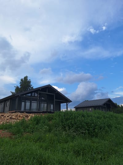 The image shows two modern, dark-colored wooden houses situated on a grassy hill under a partly cloudy blue sky. The house in the foreground is larger, with a prominent gabled roof and large windows that reflect the sky. It is built on a rocky foundation and surrounded by lush green vegetation. A tall, slender tree stands to the left of this house. The second house, smaller and further back, also features a gabled roof and is partially obscured by foliage. The environment appears rural or suburban, with no immediate signs of urban development. The scene is peaceful and natural.