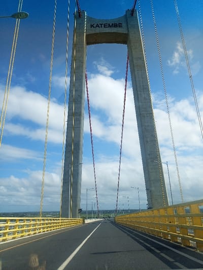 The image captures a head-on, low-angle perspective of the Maputo-Katembe Bridge, viewed from the interior of a vehicle. The scene is dominated by the bridge's massive concrete pylon structure, which features two tall, inclined supports connected by an upper crossbeam. The word "KATEMBE" is prominently displayed in dark lettering on this crossbeam.

Numerous suspension cables stretch from the top of the pylons downwards, connecting to the bridge deck. These cables vary in color, with some appearing yellow, others reddish or purplish, and some a neutral gray or white. The bridge's roadway is visible in the foreground and extends into the distance, marked by a white lane divider on dark asphalt. Bright yellow guardrails line both sides of the road, creating a strong leading line towards the vanishing point. Several streetlights are spaced along the bridge's length.

The sky above is bright blue with scattered white, fluffy cumulus clouds, indicating fair weather during the daytime. Below the bridge deck, a distant landscape of green vegetation and what appears to be a populated area is visible on both sides, suggesting the bridge spans a significant body of water or a wide expanse of land. The reflection of the vehicle's dashboard or interior is visible in the bottom left corner, confirming the vantage point. The overall impression is one of modern infrastructure against a vibrant natural backdrop in Maputo, Mozambique.