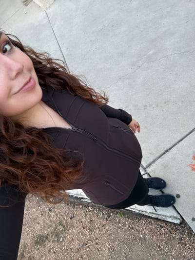 A woman stands on a sidewalk near a curb. She wears a fitted brown jacket with a zipper down the front, black leggings, and black boots. Her curly brown hair cascades around her face and down her chest. The sidewalk is concrete, and the curb is concrete and dirty. The photograph is taken from a high angle looking down at the woman.