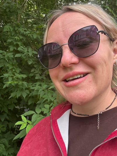 The image is a close-up selfie of a woman with blonde hair and fair skin, taken outdoors against a backdrop of lush green foliage. She is wearing large, dark-lensed sunglasses with a distinctive angular frame, through which reflections of a building with multiple windows are visible. Her mouth is slightly open, revealing her teeth and pink lipstick. She has a delicate necklace made of small black beads and a silver chain around her neck. She is also wearing a red zippered jacket over a brown shirt and a small silver hoop earring in her left ear. The lighting suggests it is daytime. The location is the city of Chastyye, Russia.