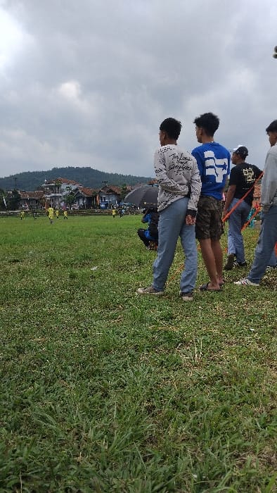 The image captures a casual outdoor gathering of young men on a grassy field in Panjalu, Indonesia, under an overcast sky. Several individuals are visible, primarily from the back or side, dressed in everyday attire.
In the foreground, a person wears a light-colored hoodie with some black text on the back, partially legible as "THE LAB," paired with blue jeans. Behind them, another individual wears a blue long-sleeved shirt featuring a large white symbol or graphic on the back, combined with camouflage shorts. Further back, a person in a black t-shirt with a white design and blue jeans is seen, with a red strap or band around their waist, hinting at participation in an outdoor game or activity. Another person in casual clothing is visible in the background. One of the individuals appears to be holding a dark umbrella.
The setting includes a wide, green grassy field, with a line of traditional-style houses with reddish-brown roofs bordering the field in the mid-ground. Beyond the houses, rolling green hills or mountains are visible under a cloudy, bright sky, indicating daytime. The overall scene suggests a relaxed outdoor event or activity.