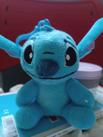A close-up, low-angle shot features a blue, plush Stitch toy sitting on top of a blue book. The toy is the primary subject, filling most of the frame. Stitch's large, black eyes with white pupils are prominent, and its mouth is a simple black stitched line. A small, darker blue nose is centered above the mouth. The toy has large, floppy ears with a hint of purple on the inside. A blue plastic loop, possibly for a keychain, is attached to the top of its head. The texture of the plush material is visible.
The Stitch toy is positioned on a bright blue book. A white barcode sticker with black text and numbers is affixed to the lower left corner of the book. The text on the sticker includes "4 00608" and "0507." To the right of the toy, out of focus, is a light pink and grey container, possibly a tin or jar. The background is a blurred, deep red surface, likely a piece of furniture, with faint dark lines suggesting a ribbed texture.
The lighting appears to be artificial, casting subtle shadows and highlighting the plush texture of the toy. There are no clear indications of the time of day or weather conditions. The setting seems to be indoors, possibly a room with red furniture. The emotion conveyed by the toy is one of simple, innocent charm. No people are visible in the image. The text "CE" is visible on the blue book, to the left of the barcode. Another line of text, "Pour les enf", is partially visible on a white surface below the blue book. The context of Ambert, France does not provide any direct visual cues within the image itself.