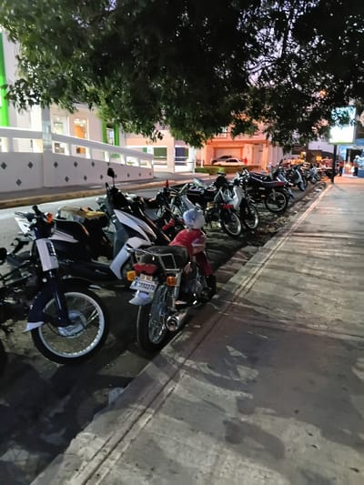The image depicts a nighttime scene in Puerto Plata, Dominican Republic, showing a row of parked motorcycles on a sidewalk alongside a building. The building features green and white accents and is partially obscured by the dark foliage of a large tree in the foreground. The lighting suggests it is dusk or early evening. Several motorcycles are visible, varying in color and style, with a prominent one in the foreground featuring a rider wearing a red jacket and a light-colored helmet. The sidewalk is made of concrete and shows some dampness or shadows. In the background, indistinct figures of people and a few cars can be seen, suggesting an urban environment. The overall atmosphere is calm, with the scene illuminated by artificial lights and the fading natural light.