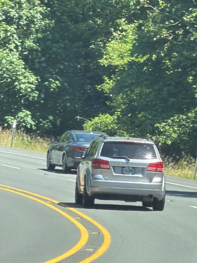 The image depicts a street scene in Seaside, United States, on a sunny day. In the foreground, a silver SUV is driving on a paved road with double yellow lines. The SUV has a license plate that appears to read "376 HFA". Behind the SUV, a dark-colored sedan is also driving on the road. The roadside is lined with dense green trees and vegetation. A white signpost is visible on the left side of the road. The overall impression is a view from a moving vehicle looking forward.