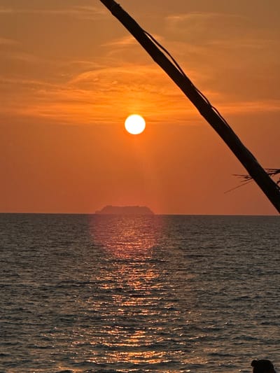 This image depicts a sunset over the sea, located in Phra Ae, Thailand. The sky is predominantly orange-red with subtle, elongated cloud formations. A bright, circular white sun disc is positioned in the upper-left quadrant, casting a prominent vertical reflection path across the dark, wavy ocean surface below. A low, dark, hazy island silhouette rests on the horizon, slightly right of the frame's horizontal center. In the foreground, a thick, dark object, consistent with a diagonal pole or tree trunk, extends from the upper-right corner downwards and towards the left. Fine, dark linear elements are visible along its surface, and dark, spiky fronds are partially visible at the bottom-right of the frame, emanating from or near the diagonal object. No human subjects or discernible actions are present.