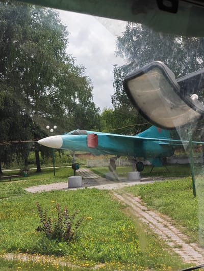 The image depicts a static display of a turquoise and white jet airplane, likely a monument or exhibit, situated outdoors in a grassy area with trees. The aircraft is positioned on a pedestal, with its nose pointed towards the left of the frame. A pathway of light-colored pavers runs through the grass. In the foreground, there is a cluster of dark-leaved bushes. The background features mature trees and a white picket fence. The sky is overcast. Reflections are visible on what appears to be a window, suggesting the photo was taken from inside a vehicle or building. The location is identified as Putyatino, Russia. There are no people visible in the image.