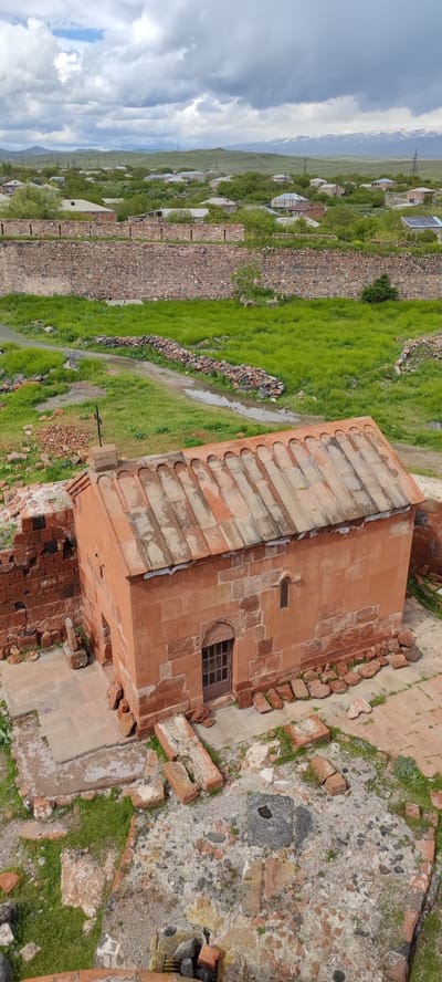 The image shows an aerial view of the Saint George Church in Dashtadem, Armenia. The church is a small, rectangular building constructed of reddish-brown stone. It has a tiled roof and a small wooden door. The church is surrounded by the ruins of other buildings and stone walls. In the background, there is a rural landscape with houses and fields. The sky is cloudy.