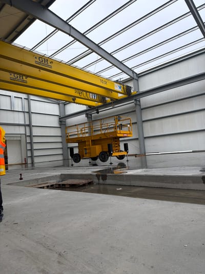 The image captures an indoor construction or industrial setting, possibly within a warehouse or factory in Jodłownik, Poland. A large yellow overhead crane, manufactured by GH Cranes, is visible suspended from the steel framework of the building. The crane has "GH" logos and "www.ghcranes.com" printed on it, along with "10.0t" indicating its lifting capacity. Beneath the crane, a yellow aerial work platform, a "H16S XL" model, is suspended mid-air, its stabilizers extended. The platform appears to be a rental unit, with the brand "renta" visible. The floor of the facility is concrete, with a section featuring a shallow pool of water and several wooden pallets. A red power drill is on the concrete floor near the edge of the water. In the foreground and left of the image, the lower body and legs of a person wearing a high-visibility orange and yellow vest are visible, suggesting their presence at the site. The building's interior features white metal siding on the walls and a translucent roof structure supported by dark grey steel beams.