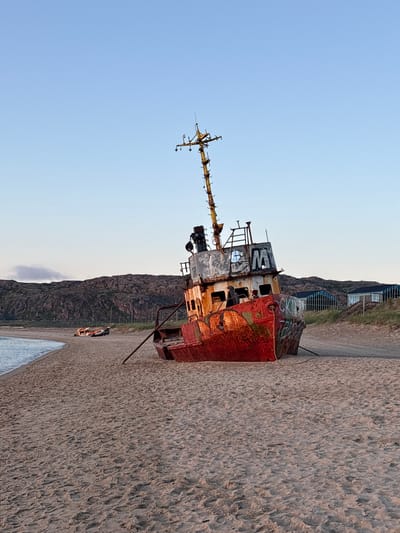 The photograph depicts a rusty, red shipwreck beached on a sandy shore in Teriberka, Russia. The ship, tilted at an angle, exhibits extensive corrosion and graffiti. Its mast and various structures are visible against a clear, pale blue sky. In the background, a rugged, rocky hillside rises, dotted with sparse vegetation. A body of water, likely the Barents Sea, laps at the shore on the left side of the image. Further down the beach, two other smaller, derelict vessels are also visible. Several glass-enclosed structures, possibly greenhouses or storage units, are situated on the slope behind the main shipwreck. The sand is textured with footprints and the overall scene conveys a sense of decay and abandonment.