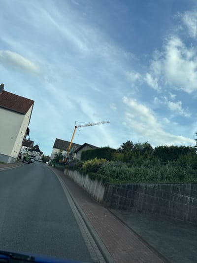 The image shows a street scene in Niederwetz, Germany, on a cloudy day. A road curves to the right, bordered by a stone wall on the left and a brick wall on the right. Dense green foliage lines the stone wall. Beyond the foliage, several houses are visible, some with red tile roofs. A yellow construction crane stands tall in the background. A green tractor is parked in front of one of the houses. The sky is partly cloudy with scattered white clouds against a blue background. The time of day appears to be daytime. No people are clearly visible, but the presence of a tractor and a construction crane suggests ongoing activity in the area. The overall impression is a typical suburban or rural setting with a touch of ongoing construction.