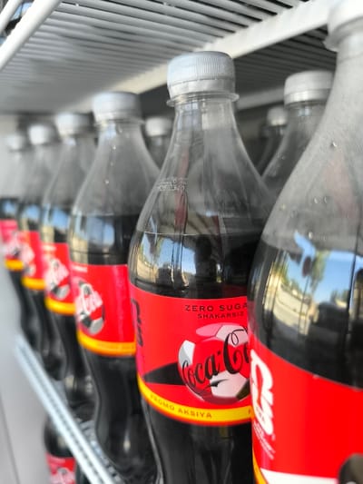 The image shows a row of Coca-Cola Zero Sugar bottles lined up on a refrigerator shelf. The bottles are plastic and have the signature red Coca-Cola label with "ZERO SUGAR SHAKARSIZ" printed on them. The label also features a graphic of a soccer ball with the Coca-Cola logo. The bottles appear to be chilled, with condensation visible on their surfaces. The background is blurred, but it suggests an indoor setting, possibly a store or a home. The location is specified as Kichik-Amirobod, Uzbekistan.