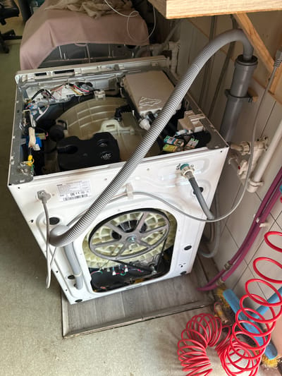 This media file shows the partially disassembled interior of a white washing machine, tilted on its side. The machine's outer casing is removed, revealing its internal components. Prominently visible is the large, white drum and its metal flywheel, driven by a motor located beneath it. Various wires and hoses are connected to different parts of the machine, suggesting maintenance or repair work is in progress. A gray corrugated hose, likely a drain or water inlet hose, runs from the top left of the machine and is connected to a pipe system above.
Other objects in the scene include red coiled hoses, possibly for air or water, a blue brush with yellow bristles, and a pink fabric item draped over the left side of the washing machine. The background shows a wooden shelf or cabinet on the left, a tiled or linoleum floor, and plumbing fixtures attached to a ceiling or wall.
The lighting appears to be indoor artificial light, and there are no strong indicators of the time of day or weather. The overall impression is of a domestic or workshop environment where a appliance is being worked on.
Visible text includes "EAC" and "CE" logos on a sticker on the side of the washing machine, indicating compliance with Eurasian and European standards respectively. There is also a model or serial number label with alphanumeric characters. The location context is Nakhabino, Russia. The image captures a moment of domestic repair or disassembly, with no specific emotional cues evident.