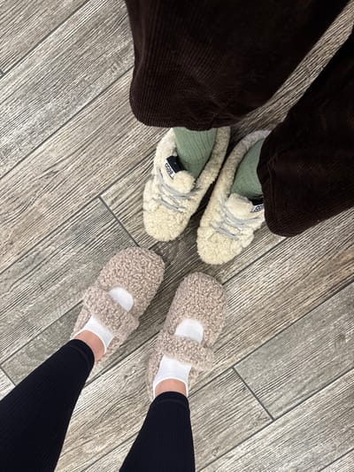 A person wearing dark corduroy pants and light-colored fuzzy shoes with sage green socks stands beside someone in black leggings and taupe fuzzy slippers with white socks. They are both standing on a gray wood-paneled floor. The shoes on the left say "GGDB" on the side. The camera angle is looking down at the floor from above.