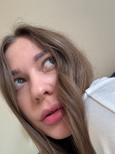 A close-up, angled portrait shows a young woman with light brown hair. The woman's face is prominent, featuring large, light-colored eyes, defined eyebrows, and full, pink lips. Part of a white garment appears at the top of the frame. The image does not provide any clues about the location or any details related to the city of Sosnovy, Russia.