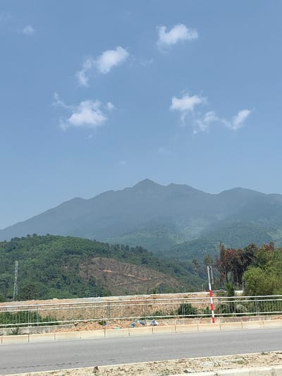 The image depicts a panoramic view of a mountainous landscape under a clear blue sky with scattered white clouds, taken in Hải Vân Ward, Vietnam. In the foreground, a paved road surface with a concrete curb is visible. Immediately beyond the curb is a gray metal guardrail, indicating an elevated structure like a bridge or overpass. Below the guardrail, a large area of excavated or leveled brown earth is present, consistent with a construction or development site. Scattered light-colored debris is visible near the base of the guardrail.
The midground features dense green foliage covering lower hills, with a distinct, lighter brown section on one hillside showing evidence of recent deforestation or earth disturbance. A tall, lattice-structured utility pole stands on the left side, partially obscured by trees. On the right, a red and white striped pole is visible behind the guardrail. The background is dominated by a range of large, hazy blue-gray mountains, their peaks rising under the bright daylight. No individuals or active movements are observed.