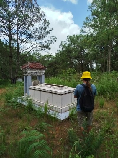 A person wearing a yellow bucket hat, a blue t-shirt, and khaki shorts stands facing away from the camera, looking at a tomb. The tomb is white and elongated, with decorative blue tile trim along the base. Above the tomb is a small, traditional roofed structure supported by grey pillars. The scene is set in a natural environment with tall green grass, ferns, and trees. The sky is blue with scattered white clouds. The person is carrying a black backpack.