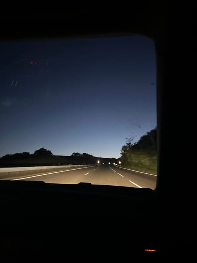 A low-angle photograph taken from inside a vehicle traveling on a road at night. The perspective is through the vehicle's window, with the frame of the window visible on the left and right edges of the image. The road is illuminated by overhead lights and the headlights of the vehicle, with green vegetation visible on both sides of the road. The sky above is dark blue. The specific location within the city of Blatino, Bulgaria cannot be determined from the image.