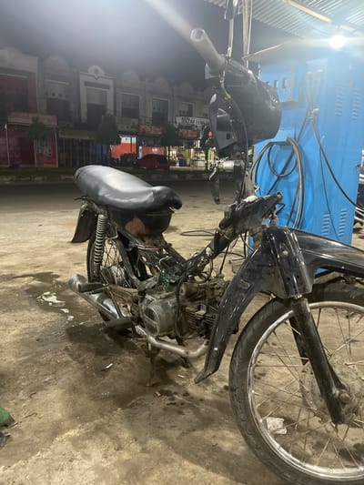 The image depicts a black motorcycle parked on a concrete ground, possibly at a gas station, at night. The motorcycle appears to be partially dismantled, with exposed engine parts and wiring. A blue structure, possibly a gas pump or a storage unit, is visible behind the motorcycle. In the background, there are buildings with shopfronts, and a red car is parked in front of one of them. The overall scene suggests an urban or semi-urban setting in Pantegajah, Indonesia. There are no people visible in the image. The ground is wet and shows signs of wear and tear, with cigarette butts scattered around. The lighting in the scene is dim, with artificial lights illuminating the area.