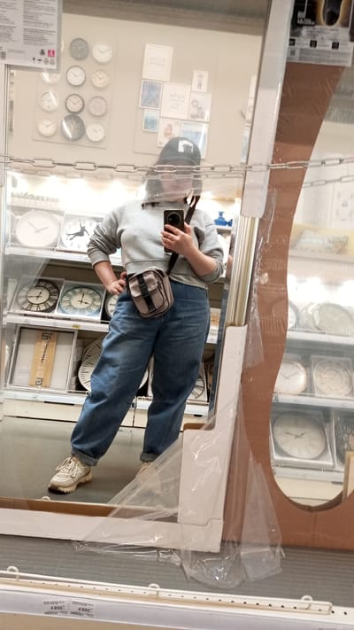 In a reflective surface, a person is shown wearing a grey hooded sweatshirt, blue jeans, and white sneakers. They also have a small crossbody bag and a black baseball cap. The person is holding a phone, presumably taking the picture of themselves in the mirror. Behind them, in the mirror's reflection, is a display of clocks and decorative items on shelves. Price tags reading "4800" are visible on a shelf below the mirror. Based on the presence of the price tags in Rubles, the location is presumably Юдино, Russia.