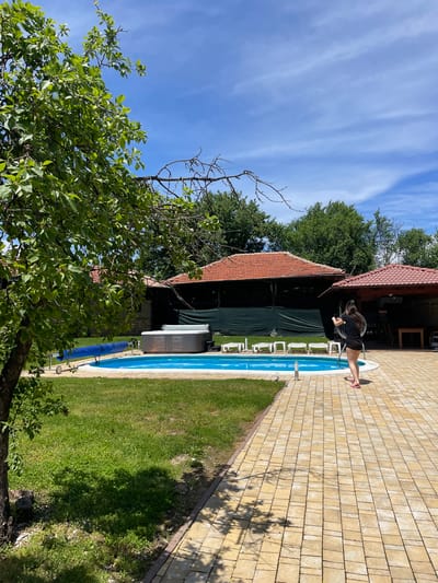 A wide shot captures a woman by a pool in Baevtsi, Bulgaria. She is standing on the edge of the pool wearing a black shirt, black shorts, and pink sandals while holding a circular object with both hands. The rectangular pool has a blue interior and white trim, surrounded by a brick patio and a patch of grass. White lounge chairs are placed alongside the pool. Adjacent to the pool is a structure with a reddish-brown roof and dark green fabric draped down. The left side of the image contains a lush tree with green leaves and bare branches extending upwards. In the background are more green trees, and a blue sky with sparse clouds.