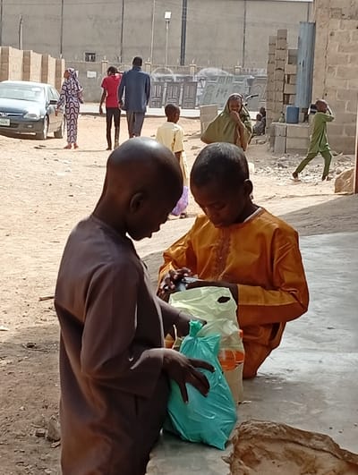 The image portrays an outdoor scene in Fanisau, Nigeria, under bright daylight, characterized by a sandy ground and informal structures. In the foreground, two young boys are the primary subjects, engrossed in examining items held within plastic bags. The boy on the left, dressed in a dark brown long-sleeved garment, is holding a green plastic bag. Next to him, a boy wearing a bright orange long-sleeved garment looks down into a light yellow plastic bag. Both boys have shaved heads and appear focused on their respective tasks.
In the midground, several other people are visible, indicating a busy environment. A young boy in a light yellow shirt stands slightly behind the two main subjects. To the right, a woman with a light green head covering and garment appears to be covering her mouth, possibly smiling or reacting to something. A boy in a vibrant green garment is captured in motion, running or quickly walking towards the right side of the frame.
The background shows more activity and elements of the setting. A silver sedan car, possibly a Honda, is parked on the left, with a partially visible license plate. A woman in a patterned blue and white outfit and head covering walks away from the car. Further back, two men are walking: one in a red t-shirt and dark pants, and another in a dark blue shirt and dark pants. The ground is unpaved and dusty. In the far background, there are concrete blocks and stacked metal gates or doors against a distant wall, suggesting a mix of residential and industrial or construction areas. The bright sun casts clear shadows, indicating midday or early afternoon. No clearly readable text is discernible.