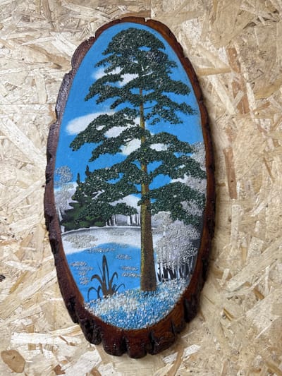 A pastoral scene is depicted in a painting mounted on a slice of wood. The painting features a tall evergreen tree as the focal point, set against a bright blue sky with scattered white clouds. The foliage of the tree is rendered with green-colored grains, creating a textured effect. In the foreground and mid-ground, the landscape appears to be covered in snow, indicated by white and light blue grains. There are dark, slender plants in the foreground, possibly reeds. In the background, a line of trees is visible, also appearing to be winter-laden. The wooden mount has a natural, rough bark edge and is placed against a background of light-colored, rough-hewn wood planks. The overall impression is a serene winter landscape, likely a decorative piece. This image does not depict any specific location within the city of Айет, Kazakhstan.