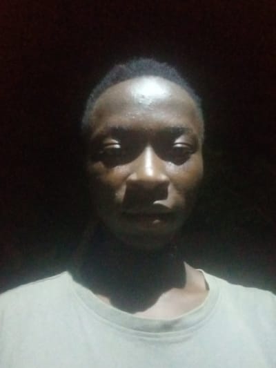 A dimly lit, close-up, front-facing selfie of a young Black man in Yola, Nigeria. The man is wearing a light-colored t-shirt. His face is illuminated from the front, creating strong shadows and highlights. The background is dark and indistinct. The lighting suggests the photograph was taken at night or in a very dark interior.