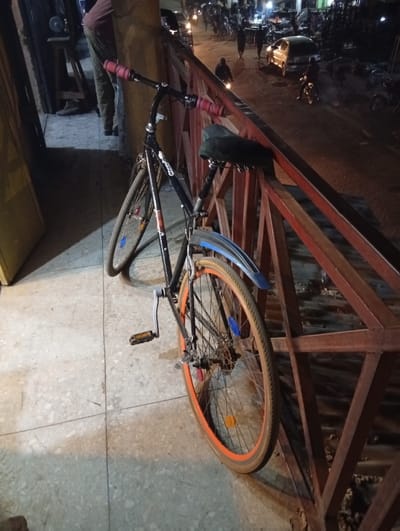A black bicycle with pink handlebar grips leans against a rusty red metal railing. The bike has orange rims and a blue fender. Beyond the railing is a street scene at night. Cars and motorcycles are visible, as are people walking. The bicycle appears to be parked on a tile surface. The overall lighting is dim, casting shadows on the ground.