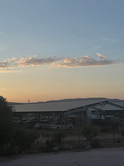 This is a horizontal, medium-shot, eye-level photograph taken at dusk. The scene is split between a vast, pale blue sky with scattered, wispy clouds that are tinged orange and pink by the setting sun, and a foreground of a building with a modern, angled roof covered in solar panels. The building appears to be a covered parking structure or a solar farm. Beyond the building, a distant mountain range is silhouetted against the twilight sky. Several vehicles, including cars and vans, are parked under the structure. A few small birds are visible in the sky, and some bushes and trees are present in the foreground. The overall mood is peaceful and serene, with a hint of industrial modernity. The image captures the transition from day to night in a somewhat arid landscape, possibly a rural or semi-urban area. The location is Rivesaltes, France.