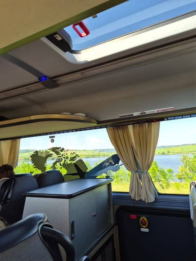 The image depicts the interior of a bus, looking out a window towards a scenic landscape. The foreground shows seats, a table/cabinet structure, and a window with a map of Canada adhered to it. The window reveals a body of water, lush green fields, and a bright blue sky with scattered clouds. The bus interior features a skylight with a red handle, interior lighting, and beige curtains. A sign with German text "PIER-NÜVE REISEN" is visible near the skylight. The lower right corner of the bus interior shows a dark panel with a red and yellow emergency button labeled "NOT-HALT." There are no discernible landmarks or features within the image that would suggest the location of Pocoleni, Romania. The scene captures a moment of travel, with the focus on the view outside and the internal elements of the bus.