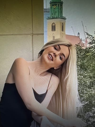 The image shows a young woman with long blonde hair, wearing a black dress. She is leaning on a railing, with her head tilted back and a smile on her face. Behind her, a historic building with a green spire can be seen, suggesting an urban environment. The woman's posture and facial expression convey a relaxed and happy mood. The lighting suggests it is daytime. The building in the background resembles the historic tower of the Saint Barbara's Church in Chorzów, Poland.