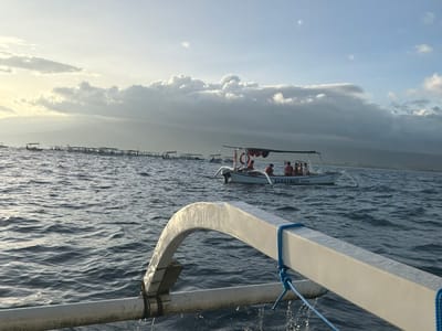 The image displays a maritime scene in the waters off Pengastulan, Indonesia. The perspective is from an active outrigger boat, with its white bow structure and visible water spray occupying the immediate foreground. In the midground, a white motorized outrigger boat, labeled with the partial text "SARASWATI Live," is depicted. This vessel features a canopy and an orange lifebuoy, carrying approximately six to seven individuals, some of whom are wearing orange life vests. In the background, a long, horizontally arrayed line of numerous smaller outrigger boats is dispersed across the choppy, dark blue water. The distant horizon reveals a hazy landmass under a partially cloudy sky, with atmospheric light indicating low sun conditions.