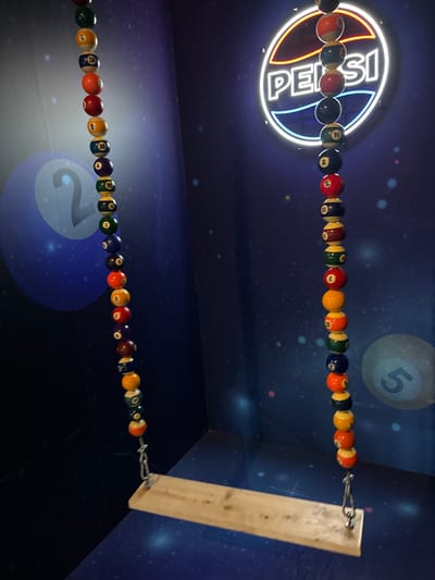 This image displays an indoor installation in the city of Arnhem, Netherlands. A swing with a plain light-toned wooden plank seat is suspended by two chains, each constructed from numerous threaded billiard balls. These balls include solids and stripes, featuring a full range of colors such as red, yellow, green, blue, black, and purple, with visible numbers including 1, 2, 3, 4, 5, 6, 7, 8, 9, 10, 11, 12, 13, 14, and 15. The balls are affixed to metal chain links that connect to the wooden seat. In the background, a circular neon sign prominently displays the Pepsi logo with a white outline, a red upper curve, and a blue lower curve, and the word 'PEPSI' in white. The environment consists of dark blue walls and a matching floor, both adorned with scattered small, bright specks, creating a cosmic or starry effect. A large, translucent light-blue number '2' is visible on the left wall, and a smaller, similar number '5' is partially visible on the right wall. No individuals are present in the scene.