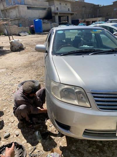 The image depicts an outdoor automotive repair scene in کوکران, Afghanistan, on a sunny day. A silver sedan, identified as a Toyota, is centered in the foreground, with its front passenger wheel removed or in the process of being serviced. A male individual, dressed in a brown traditional outfit and a patterned cap, is crouched beside the front passenger fender, actively working on the vehicle's wheel hub area with a wrench. A detached tire rests directly behind him. Various tools, loose nuts, a hubcap, and a small cardboard box with brake pads are scattered on the dirt and gravel ground.
Within the sedan, two occupants are visible in the front seats. The driver's hands are on the steering wheel, and the passenger appears to be wearing a camouflage pattern. The car's windshield displays a green circular sticker with "555 TOYOTA" and a yellow rectangular sticker labeled "F12660" with additional text in a Cyrillic-like script. A tissue box branded "ALDROZAY" is visible on the dashboard.
The background features multiple unpainted, multi-story brick and concrete structures, appearing to be residential or commercial buildings. One building has a large blue water tank positioned next to it, and a prominent dark, possibly soot, stain on its upper wall. The ground in the background is also uneven dirt, with some scattered debris and what appears to be discarded corrugated metal sheets. The sky is clear and light blue.