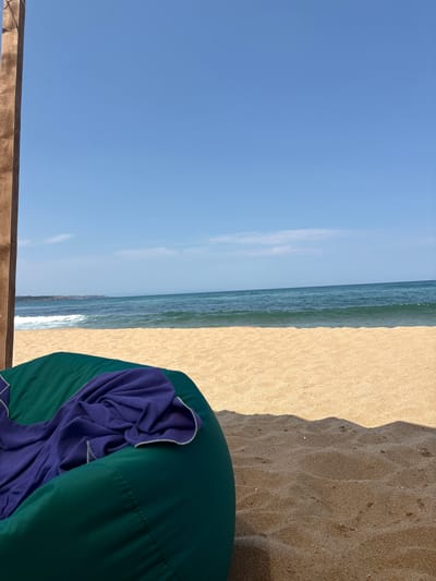 A green beanbag chair with a purple blanket draped over it sits on a sandy beach. The beanbag is in the foreground, to the left of the frame. The beach is composed of light brown sand with some textured ripples. In the background, the blue sea meets the horizon under a clear blue sky. A wooden post is visible on the far left, partially framing the scene. The location is the beach in Dyuni, Bulgaria, a resort town on the Black Sea coast.