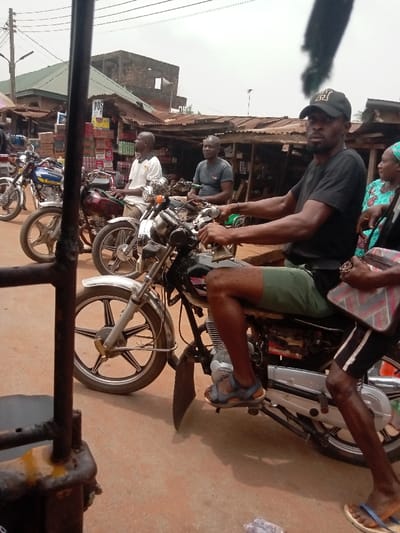 The image depicts a street scene in Umudioka, Nigeria, under an overcast sky. In the foreground, a man with dark skin, wearing a black t-shirt, olive green shorts, black baseball cap with a white 'H' logo, and blue sandals, sits astride a black motorcycle with chrome accents and alloy wheels. He holds the handlebars and looks directly towards the viewer, his left foot on the ground. To his immediate right, a portion of another individual, possibly a woman, wearing a teal patterned garment, is visible. Further right, a person's lower legs and feet in blue flip-flops are partially visible.
In the midground, two additional men of similar complexion are seated on motorcycles. The man on the left wears a white t-shirt and is on a burgundy motorcycle with spoke wheels. The man to his right wears a dark grey t-shirt and is on a darker motorcycle. Other motorcycles are visible in the background, parked or stationary. The ground surface is reddish-brown dirt.
The background features a row of market stalls with corrugated metal roofs, displaying various stacked goods and packages. A white sign with "POST" in black lettering is visible on one stall. Behind the stalls, a weathered multi-story building with exposed brickwork stands under the distant skyline. A dark, metal structural element partially frames the left foreground.