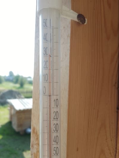 A thermometer is attached to a wooden structure in Батово, Russia. The thermometer, which is partially obscured by a white mounting bracket, displays a temperature of approximately 25 degrees Celsius, indicated by the red liquid level. The background is blurred but shows greenery, a wooden structure resembling a shed or small building, and a fence. The wooden structure to which the thermometer is attached appears to be made of light-colored wood with visible grain. The lighting suggests it is daytime with sunlight.