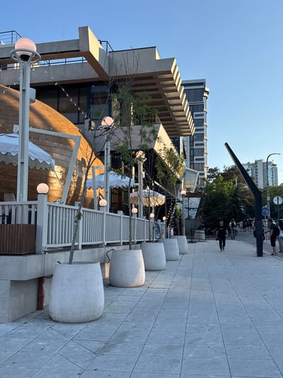 The image depicts a street scene in Neberdzhaevskaya, Russia, under a clear blue sky. The foreground features a paved sidewalk with a row of large, white planters containing trees. To the left of the sidewalk, a white fence separates it from an outdoor seating area with white umbrellas. Behind the fence, a curved wooden structure with windows and lights is visible, suggesting a restaurant or commercial establishment. Further back, a modern building with multiple stories and glass windows rises into the sky. Several people are walking on the sidewalk, some in groups and others individually. The overall impression is of a bustling urban environment with commercial activity and pedestrian traffic.