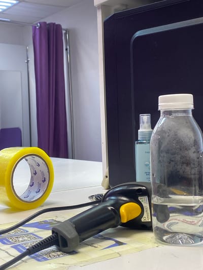 The image depicts an indoor setting, likely a retail or service environment, with a focus on several objects. Dominating the foreground and midground is a black and yellow barcode scanner, connected by a coiled cable. Next to the scanner is a roll of yellow Scotch tape, with visible branding. Behind these items, a clear plastic bottle with a white cap is positioned, containing a liquid with particles. Reflected in the bottle is a spray bottle with blue labeling and text.
The background shows a dark surface, possibly a piece of equipment or furniture, and a section of purple fabric, likely a curtain or drape, hanging from a metal rod. The overall lighting suggests an indoor environment, with no clear indication of the time of day. The scene is static, with no active subjects or events taking place.
Visible text includes "Scotch" multiple times on the tape, and brand or model information on the scanner, such as "MINDEO," "KKO1204212," and "MADE IN CHINA," along with a "CE" mark. There are also Cyrillic characters visible on the spray bottle and on some paper or stickers in the background. The location context provided is Uchaly, Russia, which may align with the Cyrillic text observed.