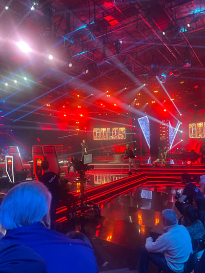 A stage setup with red lighting and beams shines brightly. Camera operators and audience members are present, partially obscuring the view. A metal structure with lights is visible on the left side. The word "GLUE" is prominently displayed in large, lit-up block letters. Red accents and beams run throughout the stage's design. The stage appears to be set up for a show or performance.