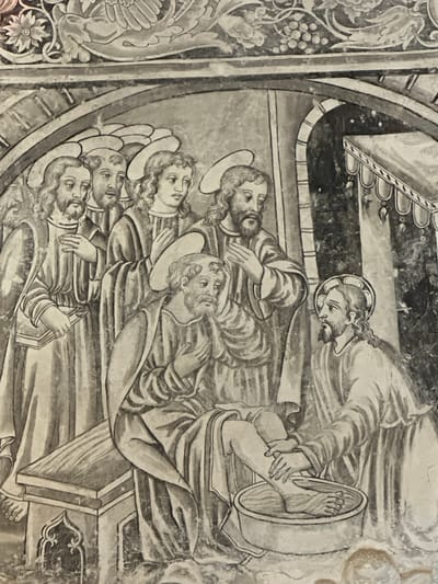 This is a fresco depicting Jesus washing the feet of Saint Peter. Jesus, with a halo, is kneeling and holding Peter's foot, which is in a basin of water. Peter, also with a halo, is sitting and looking down at his foot with an expression of surprise or humility. Around them stand other figures, presumably apostles, also with halos, dressed in robes. The scene takes place within an architectural setting that includes an archway and a curtained alcove. The fresco is located in the Augustinian convent of San Miguel in Huejotzingo, Puebla, Mexico.