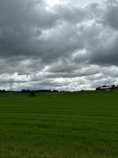 A wide shot captures a stormy sky over a green field in Kråkstad, Norway. The sky is dominated by dark, heavy clouds, with patches of lighter gray suggesting breaks or thinner areas. In the middle ground, a bright green field stretches horizontally across the frame, with visible rows from farming practices. A silver electrical pylon stands tall on the left side of the field, with power lines extending across the scene. In the distance, on a gentle slope to the right, a red barn with a white roof and trim is visible, accompanied by a few scattered trees and smaller structures. The overall impression is one of an approaching storm in a rural landscape.