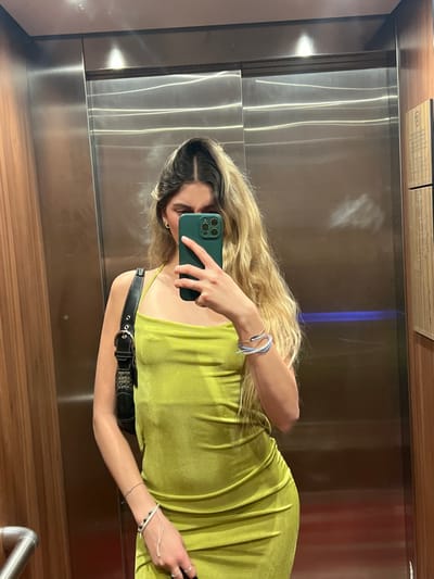 The image shows a young woman standing inside an elevator in Val-d'Isère, France. The woman is wearing a lime green, form-fitting dress and a black shoulder bag. She is holding a green smartphone in front of her face. The elevator walls are made of metal, and the ceiling has recessed lighting. The woman's right arm is extended in front of her, and she is taking a selfie with her phone. The background of the image is the metal interior of the elevator. The woman's left arm is bent at the elbow, and her hand is resting on her hip. She is wearing a small silver bracelet on her left wrist. The image is lit from above. The elevator is moving.